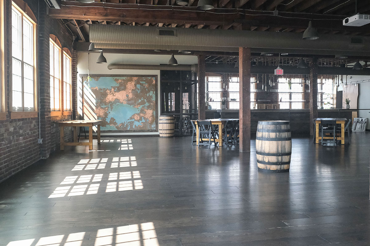 Photo of an open industrial inspired event space. Brick exterior walls with tall sun filled windows that stretch up to a high open ceiling with exposed wood beam rafters. A large teal and copper colored abstract painting covers the far wall next to a long bar separated from the space by open shelving. Large barrels and a few tables with chairs are scattered around the dark wood floor.