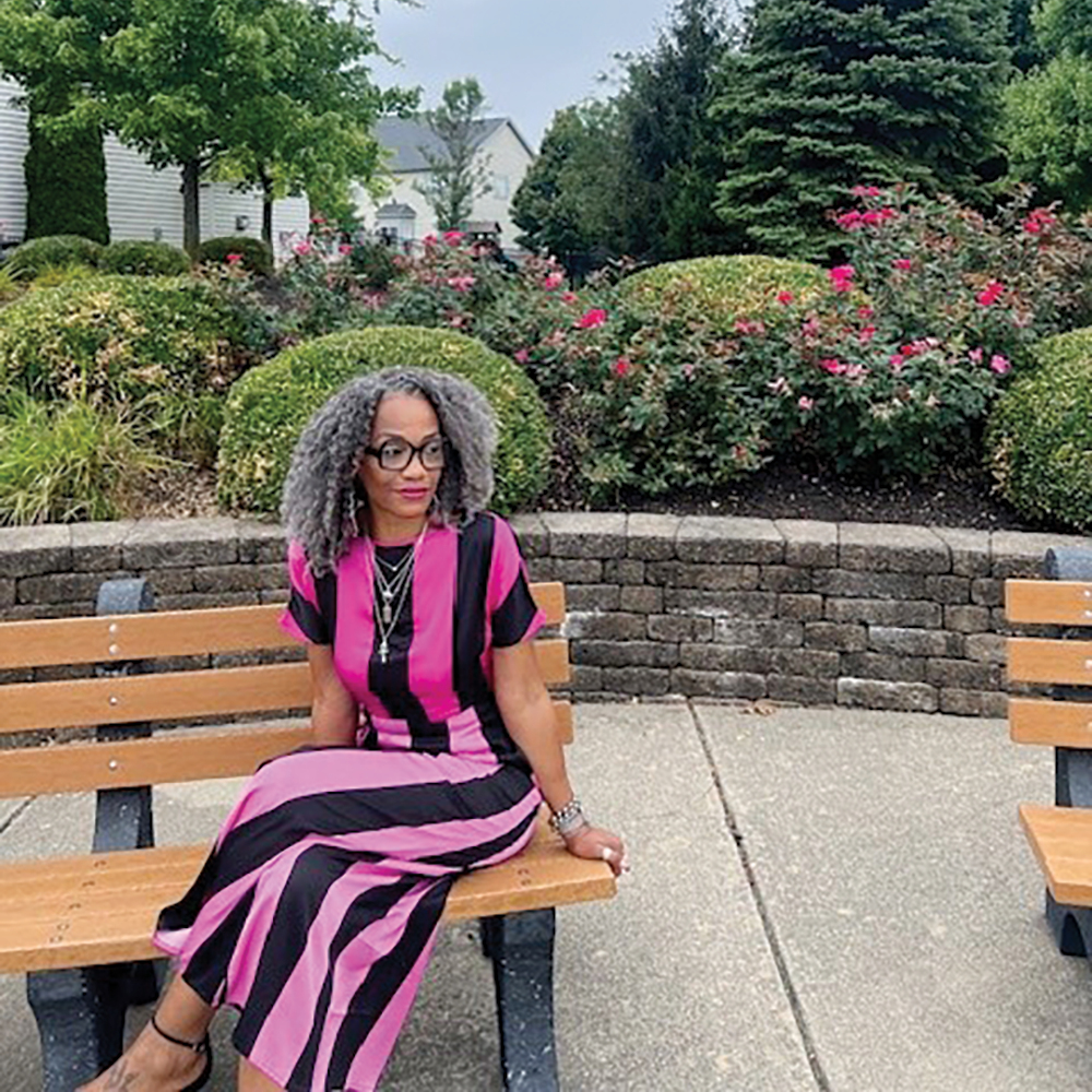 A photograph of Alicia West sitting on a bench outside in front of a rose and hedge filled garden bed wearing a pink and black striped outfit