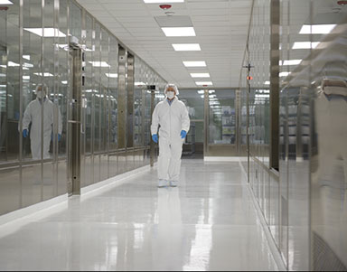 A lone individual in laboratory ppe including white scrubs, hair cover, face mask, and blue latex gloves walks down a brightly lit sterile looking hallway with mirrored walls