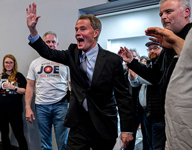 Indianapolis mayor Joe Hogsett, dressed in a suit, stands at the center of frame with a hand in the air and an open mouthed smile surrounded by celebrating onlookers