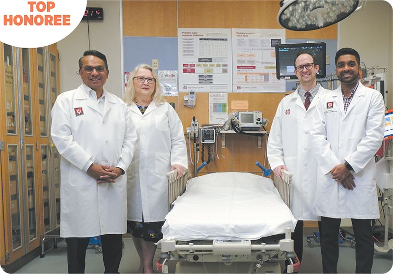 Group photograph of Dr. Mitesh Shah; Dr. Laurie Ackerman; Dr. Matthew Partain and Dr. Satyan Sreenath standing around a bed in a hospital room, in the top left corner text reads Top Honoree