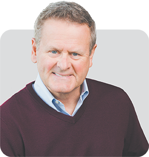 A short haired man wearing a wine colored sweater over a light blue collared shirt smiles at the camera