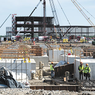 A construction site with a poured concrete foundation and steel framing in the distance. Many workers in hard hats and vests stand around the site.