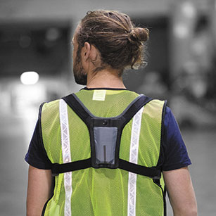 A bearded man with long hair in a bun stands in a warehouse with his back to camera wearing a high visibility vest and a device harness