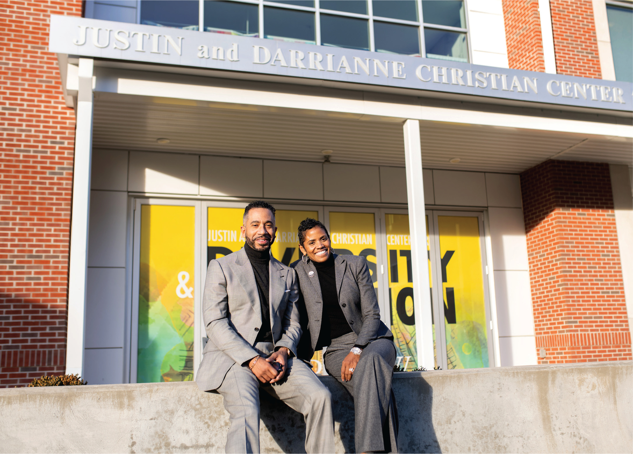Justin and Darrianne Christian, a black couple both wearing grey suitsover black turtlenecks, sit in front of a building's entrance under a sign reading Justin and Darrianne Christian Center