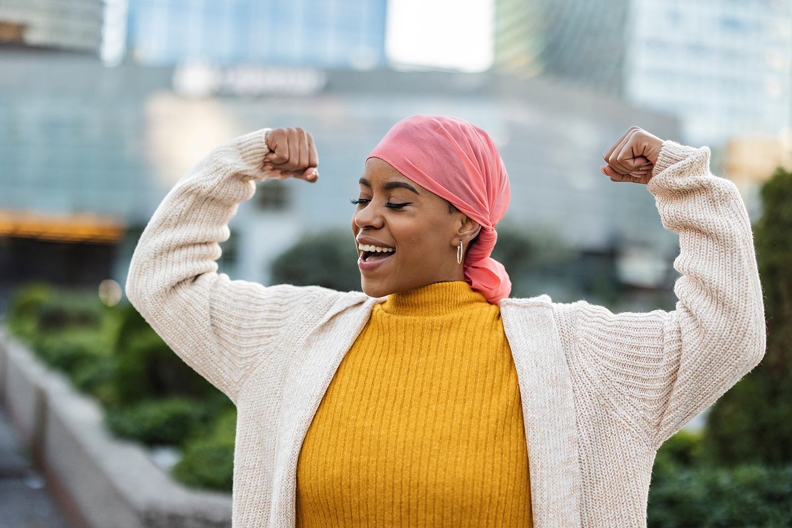 A black woman wearing a pink scarf over her hair and chunky neutral tone knits smiles and poses with her arms curled in a double bicep pose.