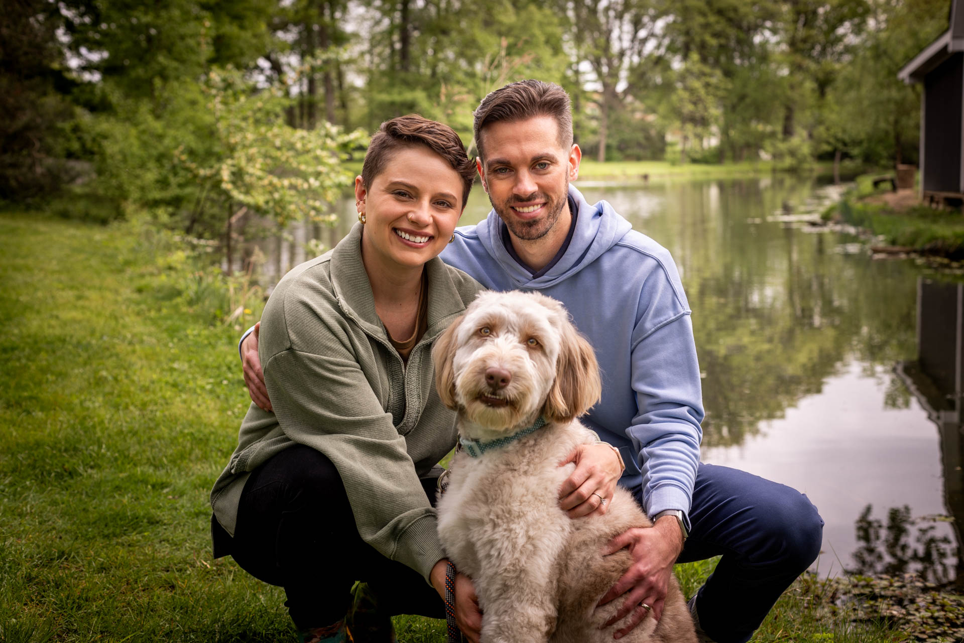 A man and woman crouch down in the cut grass next to creek with their hands on the white dog in front of them.