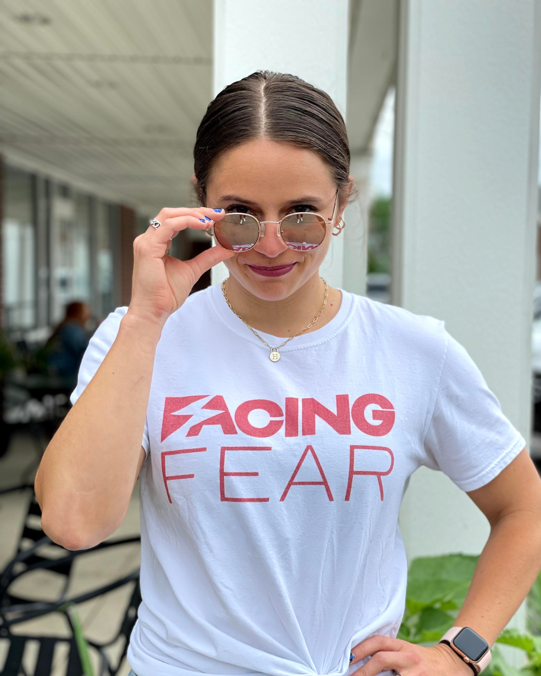 A woman with hair pulled back in a center part wearing a T shirt that reads Facing Fear pulls her round framed mirror lens sunglasses down her nose and looks over the lens at the camera.