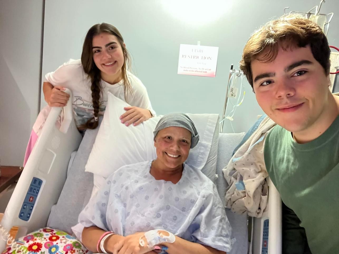 A woman in a hospital gown sits up in a hospital bed, her daughter stands behind her left shoulder and her son is in the foreground to her left. All three smile at the camera