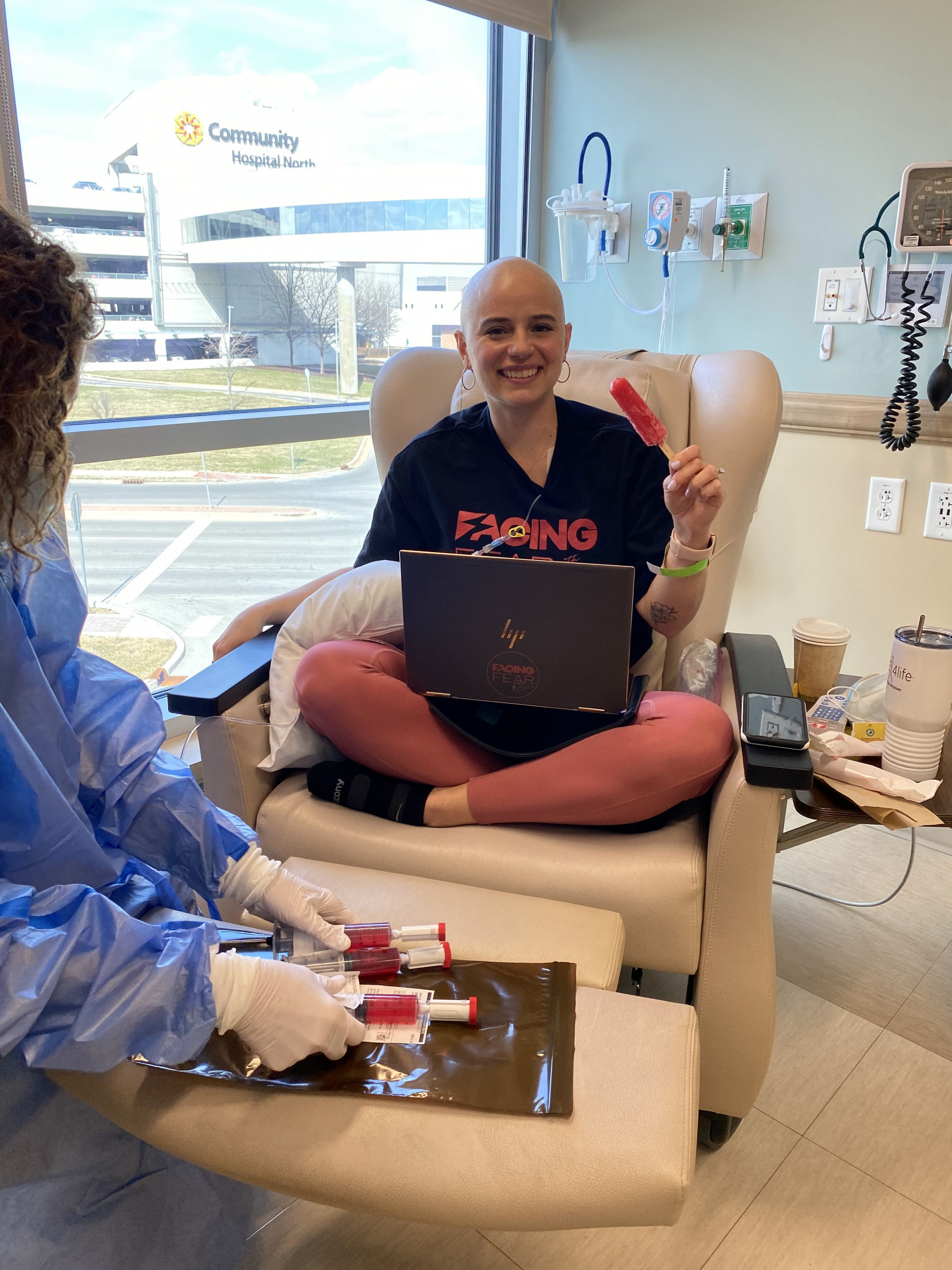 A woman seated in the chair of a hospital room smiles at the camera with a laptop on her crossed legs and a popsicle in her hand