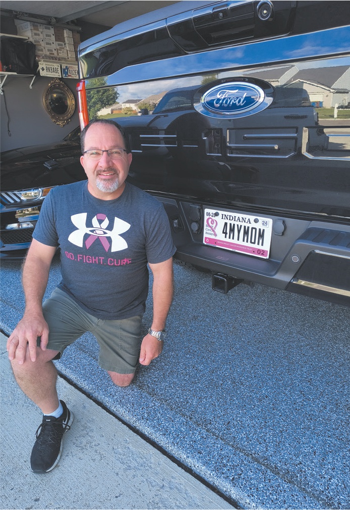 A man in a Breast Cancer Awareness branded t shirt poses in front of his vehicle's Breast Cancer Awareness license plate