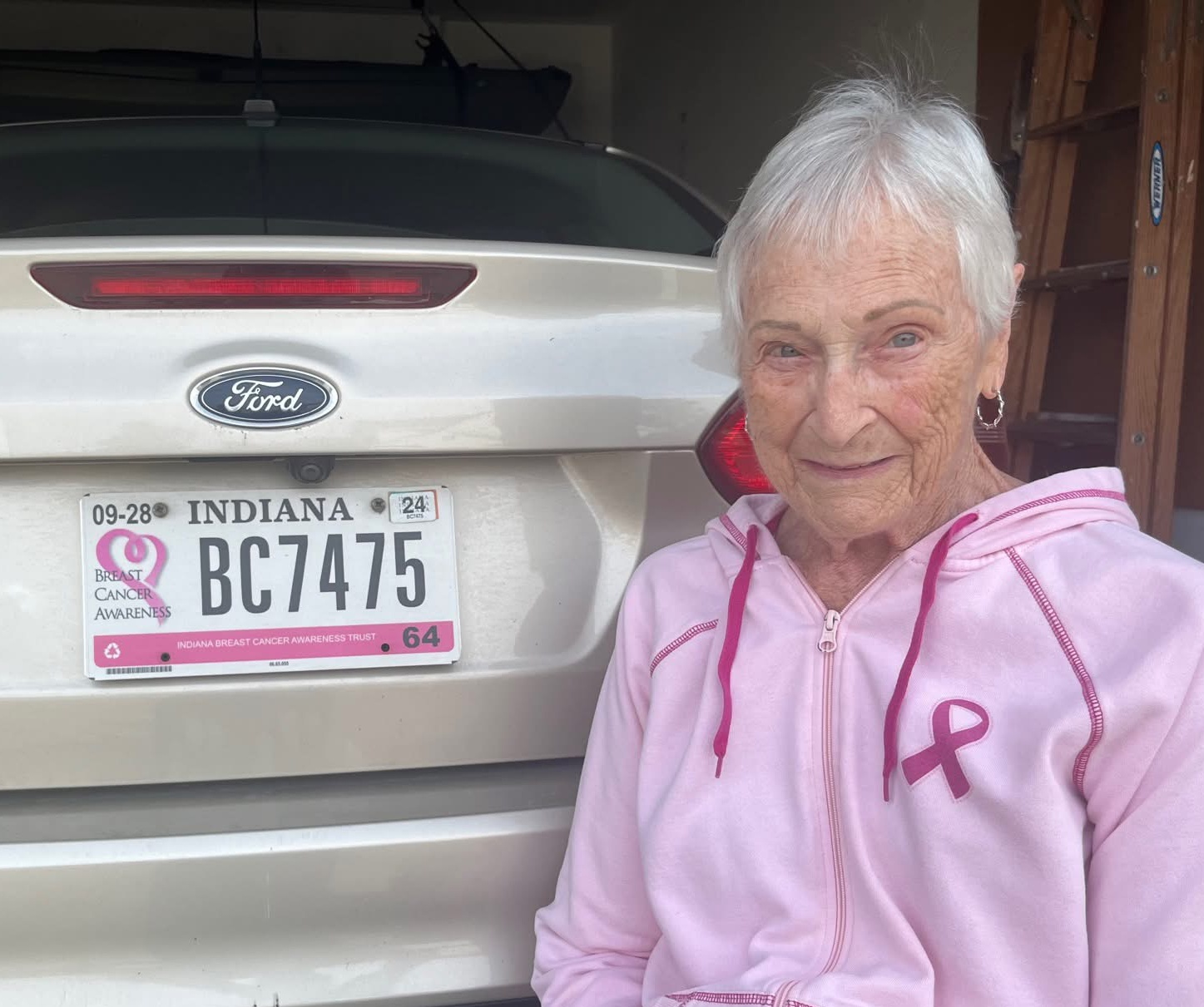 A woman in a pink zip up hoodie poses in front of her vehicle's license plate. Her sweatshirt and license plate both feature the pink breast cancer awareness ribbon.