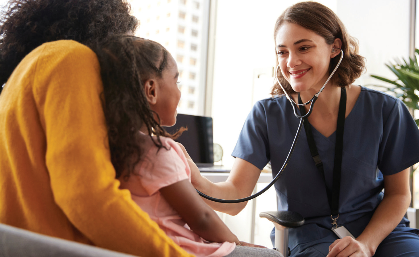 A smiling medical professional holds a stethoscope to a child in her mother's arms.