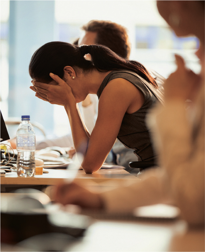 Photograph of a woman hunched head hanging over a communal desk with her face in her hands