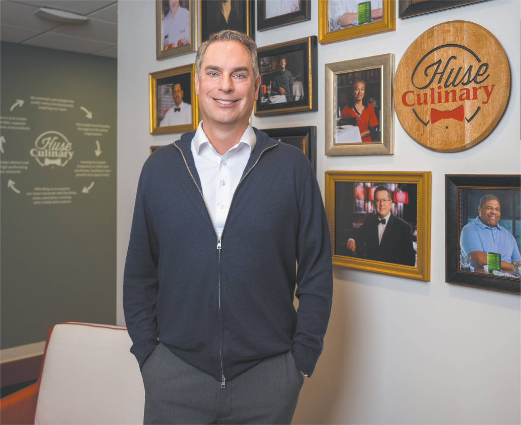 Craig Huse smiling in front of a wall of photographs with a Huse Culinary plaque